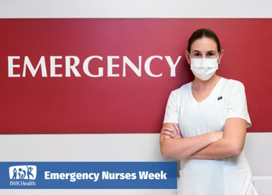 Nurse wearing mask with arms crossed stands in front of the word emergency painted white on a red wall.