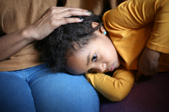 A dark skinned girl rests her head in her mother's lap and holds her tummy.