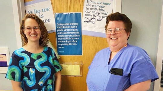 A woman in a black shirt with thick blue and green squiggles on it stand to the left of a woman in nurse scrubs. They both are smiling widely.