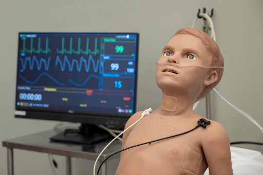Simulation 'dummy' of child sits up in bed with health monitoring equipment beside it.