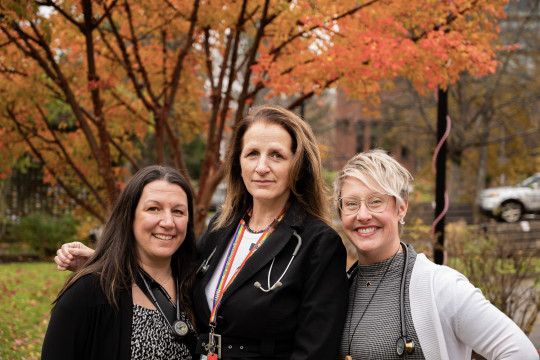 Three nurse practitioners stand outside in front of a tree with autumn foliage