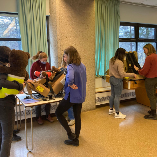 Participants in a Child Safety Link training session practice using child car seats with safety equipment and dolls. They are gathered in small groups, actively engaged in hands-on learning.