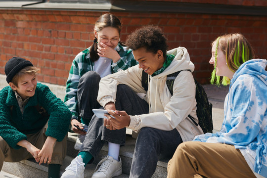 A group of teens sit on the steps laughing and looking at a phone screen.