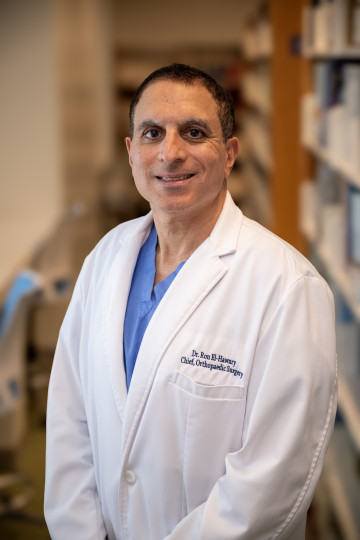 Man in lab coat standing in library wearing a lab coat which reads Dr. Ron El-Hawary, Chief Orthopaedic surgery.