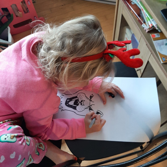 A little girl wearing red reindeer antlers leans over a picture she is drawing of a lion.