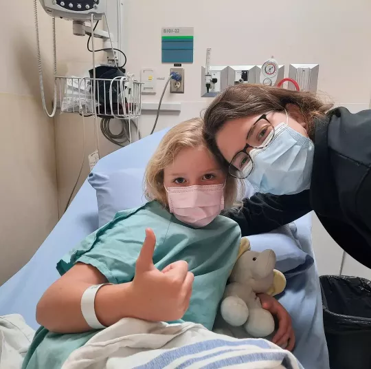 A little girl in a mask and hospital bed gives a thumbs up as a caregiver leans in to be in the picture.