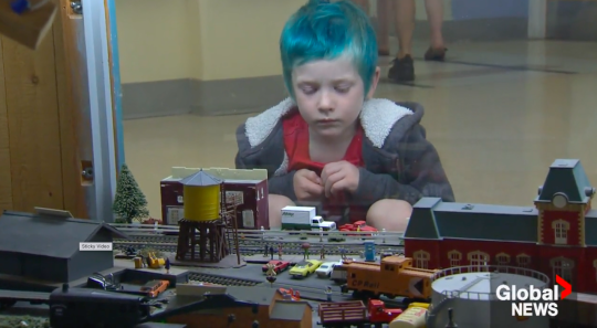 A child with dyed green hair looks at a model railroad through plexiglass.