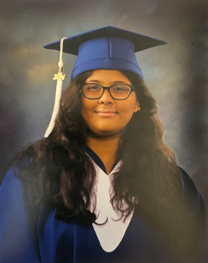 Young woman with light brown skin and glasses poses in grad cap and gown.