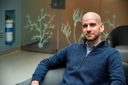 Man in blue zip up sweater sits in front of glass wall etched with seaweed motif.