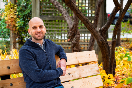 Man in blue zip up sweater sits ion bench in front of wooden pergola with yellow autumn leaves on the ground.