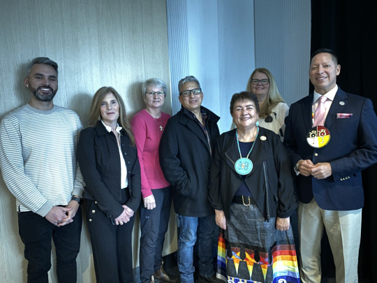 Seven people stand together, two wearing indigenous accessories,