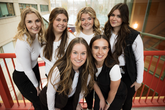 Six Nurses in white long sleeve tees and black vests stand together on a bridge overlooking an atrium