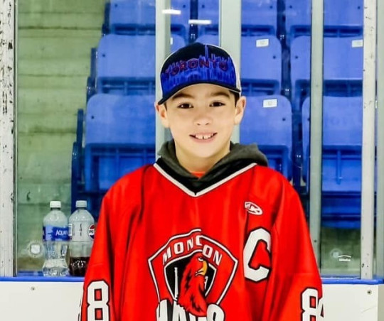 A boy in a baseball cap and hockey sweater stands in a rink.