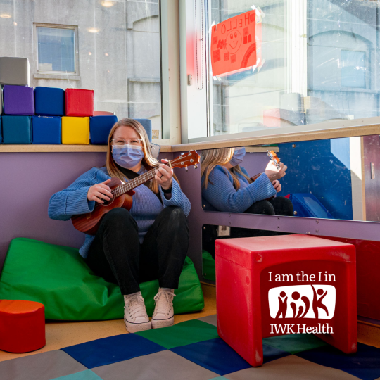 A woman sits on a big cushion in a playroom, a ukulele in her hands.