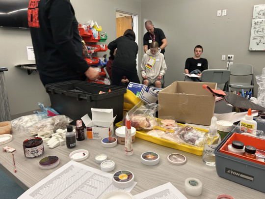 Table of costume make up with simulation volunteers in the background having fake injuries applied