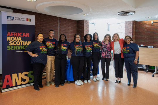 Nine people, six of African Nova Scotia descent, stand in front of a banner that reads African Nova Scotian Services. Their t-shirts and banner are dark blue with red, green and yellow lettering.