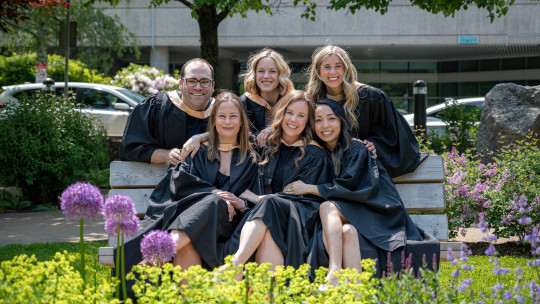 Six smiling grads with big smiles gather around a park bench.