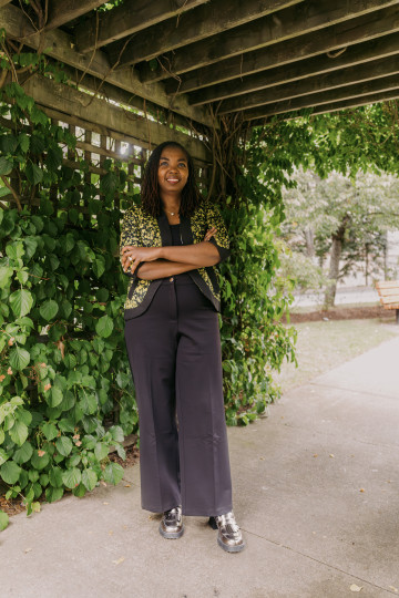 Afolake Awoyiga, wearing black pants and gold jacket, stands under a pergola.