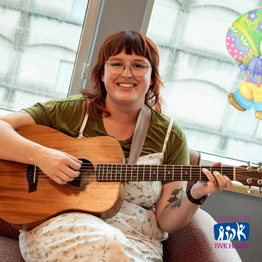 Sarah Bourque sits in a playroom holding a guitar.