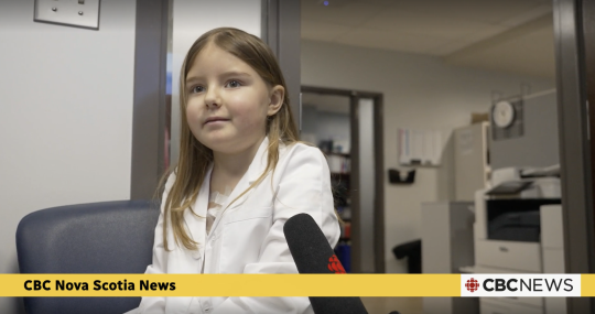 A young female patient speaks into a CBC news microphone. She wears a lab coat.