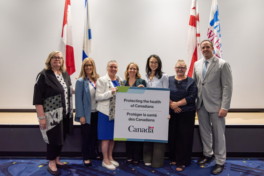 Seven dignitaries stand holding up a sign that says Protecting the Health of Canadians.