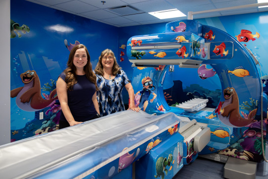Nicole Mroz and Sandra Ellis stand by a CT scanner decorated with friendly cartoon sea creatures