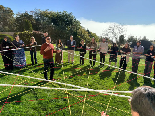 John Sylliboy stands in the middle of a dreamcatcher created by people in a circle holding threads connected to one another.