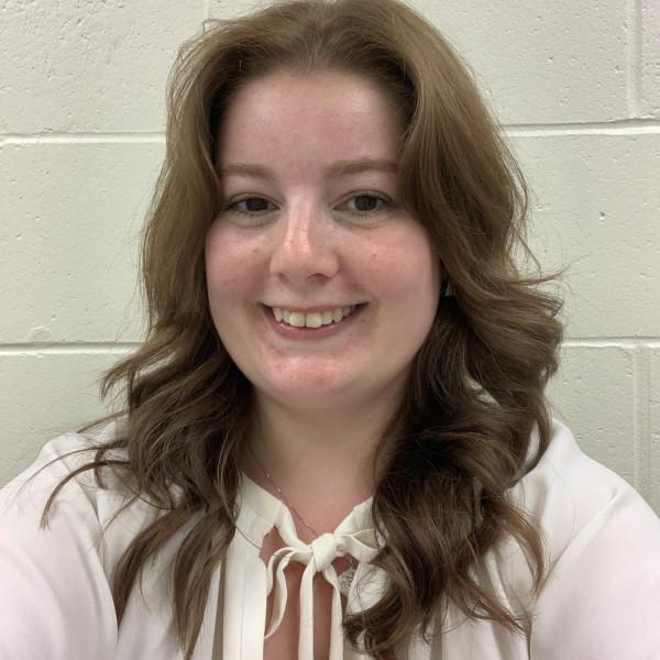 A woman with shoulder-length wavy brown hair smiles at the camera. She is wearing a light-colored blouse with a bow at the neckline, standing in front of a white brick wall.