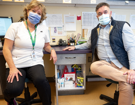Woman in white shirt and man with blue shirt and dark blue vest sit on either side of an open cabinet. Inside the cabinet are an array of products to ease pain. Both people are masked.