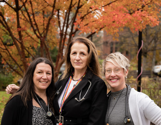 Three nurse practitioners stand outside in front of a tree with autumn foliage
