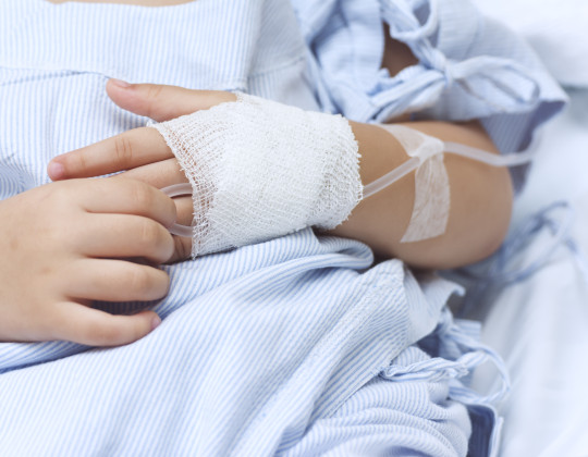Close-up of a child's hands are clasped in front of them. They wear a hospital gown and have an IV running to a bandage on one hand.
