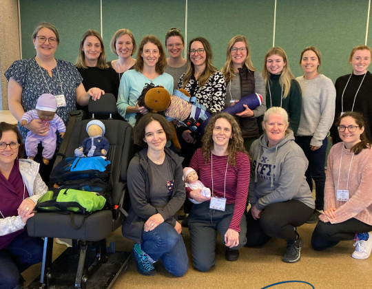 Group of women smiling and posing with child safety equipment and dolls at a Child Safety Link training event. Some are holding dolls, while others are gathered around a car seat simulator. The Child Safety Link logo is visible in the bottom right corner.