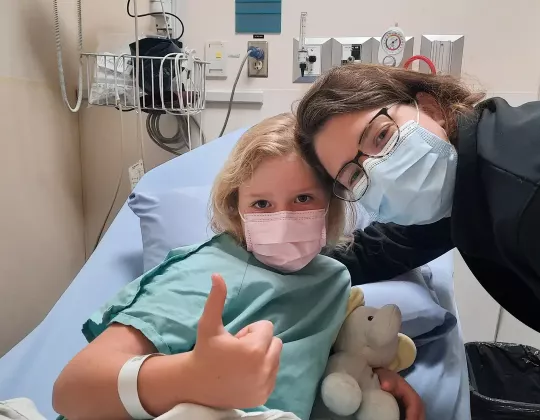 A little girl in a mask and hospital bed gives a thumbs up as a caregiver leans in to be in the picture.