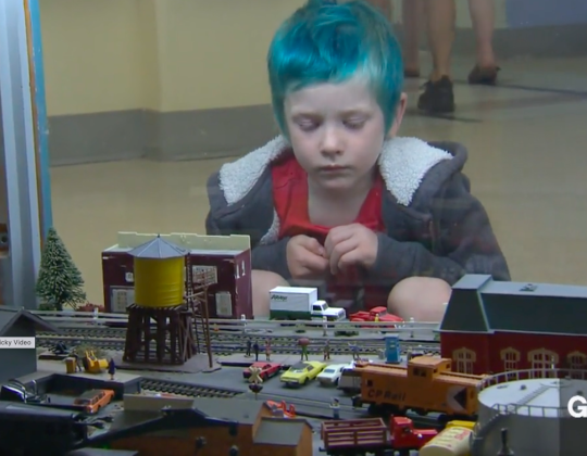 A child with dyed green hair looks at a model railroad through plexiglass.