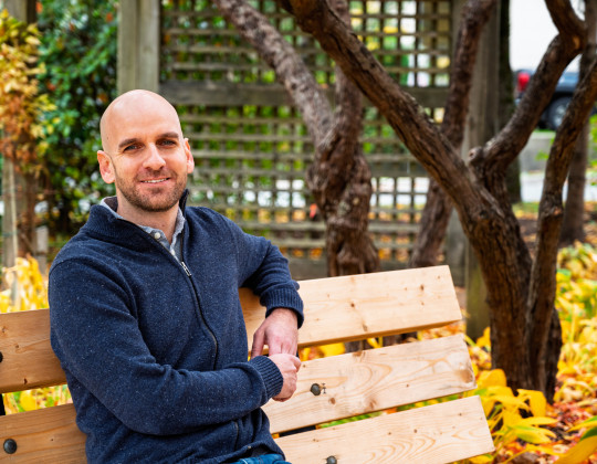 Man in blue zip up sweater sits ion bench in front of wooden pergola with yellow autumn leaves on the ground.