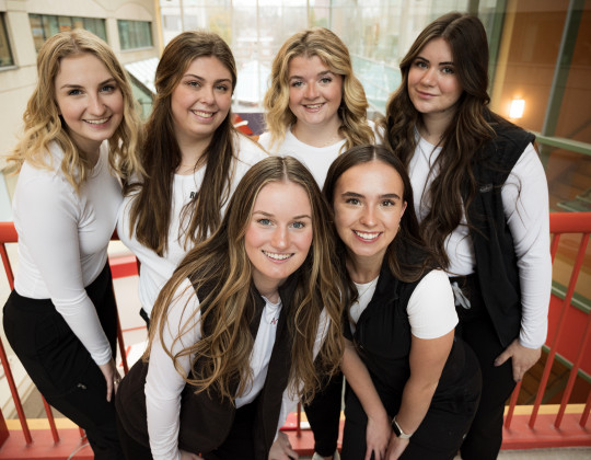 Six Nurses in white long sleeve tees and black vests stand together on a bridge overlooking an atrium