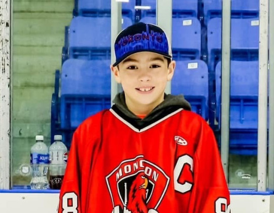 A boy in a baseball cap and hockey sweater stands in a rink.