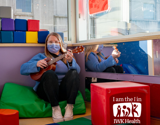 A woman sits on a big cushion in a playroom, a ukulele in her hands.