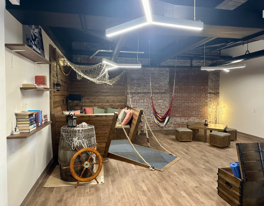 A comfortable space for youth with a booth that looks like the prow of a boat and a swing seat hanging from the ceiling. There are shelves of books on the wall and a ship's steering wheel leans against a barrel.