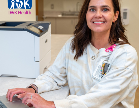 Jill Forbes smiles as she types on a laptop. There is an I am the I in IWK logo in the corner of the photo.