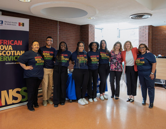 Nine people, six of African Nova Scotia descent, stand in front of a banner that reads African Nova Scotian Services. Their t-shirts and banner are dark blue with red, green and yellow lettering.