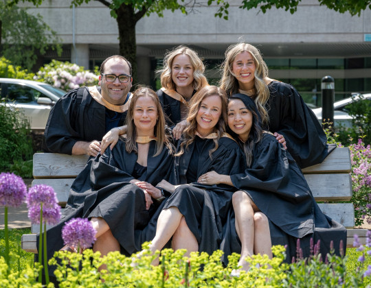 Six smiling grads with big smiles gather around a park bench.