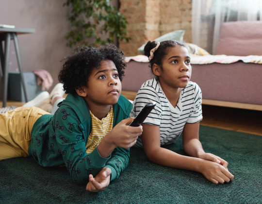 Two African Canadian children lying on floor and watching TV together during leisure time at home.