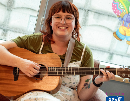 Sarah Bourque sits in a playroom holding a guitar.