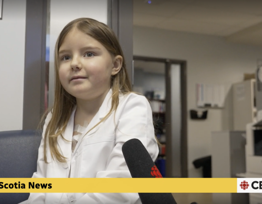 A young female patient speaks into a CBC news microphone. She wears a lab coat.