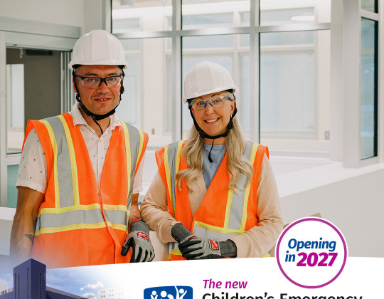 Brian and Katie stand in the under construction Emergency department wearing safety vests, helmets and goggles.