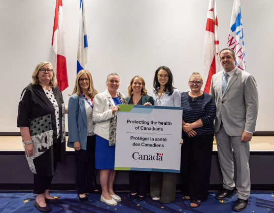 Seven dignitaries stand holding up a sign that says Protecting the Health of Canadians.