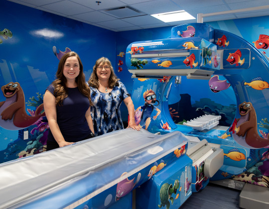 Kaitlyn Carvery and Sandra Ellis stand by a CT scanner decorated with friendly cartoon sea creatures