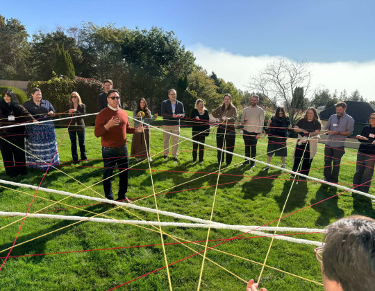John Sylliboy stands in the middle of a dreamcatcher created by people in a circle holding threads connected to one another.
